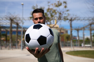 Handsome young man holds a football in his hand and shows it to the camera in focus. The man is a...