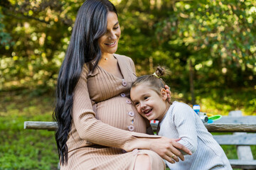 Happy pregnant mother and daughter hugging on a park bench. Happy motherhood. Beautiful woman with a little daughter in nature