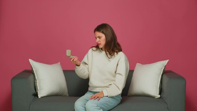 Dissatisfied young girl holds gift box with engagement ring unpleased with marriage proposal, grimacing, sitting on sofa