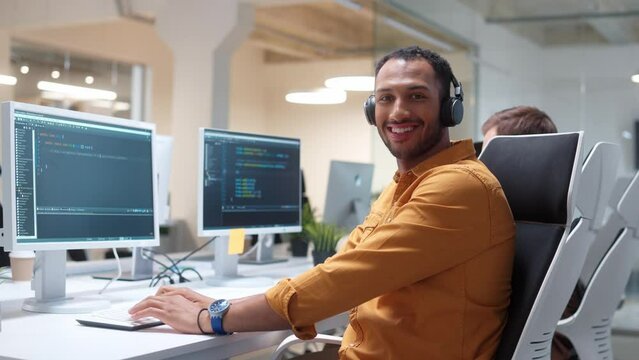 Camera approaching young Hindu man IT programer working on desktop computers typing on keyboard coding and listening to music in headphones. Close up of professional worker in modern office