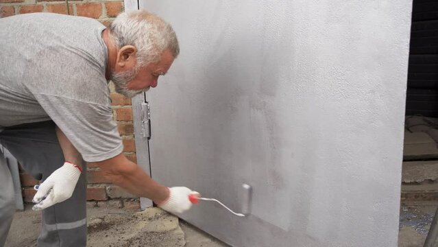 an elderly gray-haired man paints the metal garage door with a roller. 