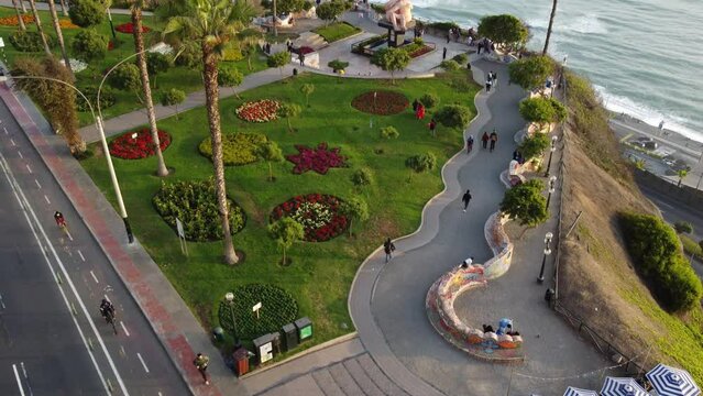 Drone 4k video of a park of a park on edge of cliff in Lima Peru in Miraflores. Drone tilts up. Below are palm trees, people walking, grass, and below is the pacific ocean beach.