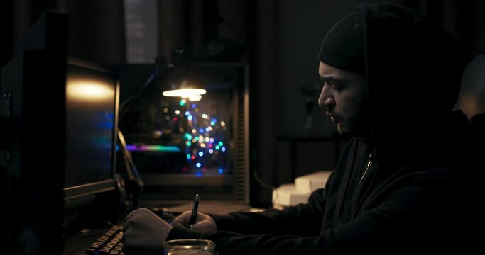 Man in black jacket sits at computer desk in dark room. Guy with beard holds pen in hands and takes notes. In front of him are monitors on which files are loaded. The hacker is working on task.