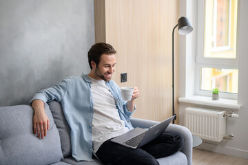 Serene young 30s caucasian guy using laptop in relaxed home atmosphere, sitting on the sofa and typing, web surfing, watching movie. Carefree man drinking beverage
