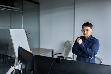 Serious and thinking asian engineer looking at two monitors, close-up photo of office worker, man working inside modern office building, businessman wearing glasses and casual shirt.