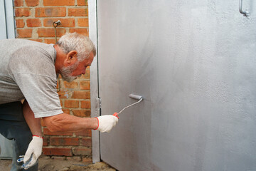 an elderly gray-haired man paints the metal garage door with a roller. 