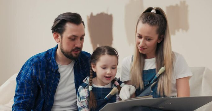 8-year-old girl dressed in denim overalls and checkered shirt unhappily reviews plan for future renovation of apartment with parents. The child would like future room to be more spacious.