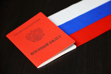 Russian military ID and national flag on wooden table. Translation of inscription: Military ID card, Russian Federation. Concept of mobilization in Russia