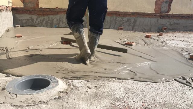 Construction worker laying lightweight concrete on a roof top in a construction site. Man with boots expanding cement on a building roof with a drain in the shot. Worker making slopes on the deck.