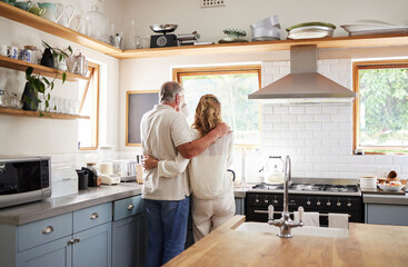 Couple hug, morning coffee and senior people having breakfast in the kitchen while looking at outdoor nature. Elderly man and woman hugging with tea drink in house with love together from behind
