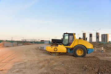 Obraz premium Soil Compactor leveling ground at construction site. Vibration single-cylinder road roller on construction road. Road work and asphalt laying. Tower cranes build high-rise buildings. Road construction
