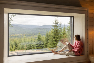 Woman sits with phone on window sill and enjoys scenic view on mountains while resting in wooden house on nature. Recreation and escaping to nature concept