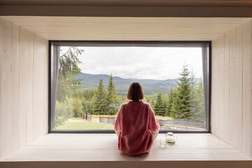 Woman meditates while sitting on window sill and enjoys panoramic view on mountains. Recreation and...