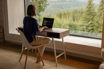 Woman works on laptop while sitting by the table in front of panoramic window with great view on mountains. Remote work and escaping to nature concept