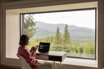 Woman works on laptop while sitting by the table in front of panoramic window with great view on mountains. Remote work and escaping to nature concept