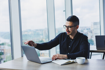 Young successful male worker in glasses and shirt sitting at desk and working on project via laptop in modern office looking at screen