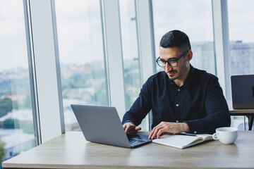 Young successful male worker in glasses and shirt sitting at desk and working on project via laptop in modern office looking at screen