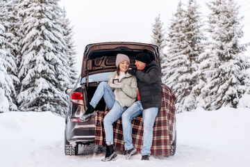 Young couple in love sitting in an embrace in the open trunk of a car,drinking tea in a winter forest.Winter activities,active lifestyle,Valentine's day,tenderness and love.