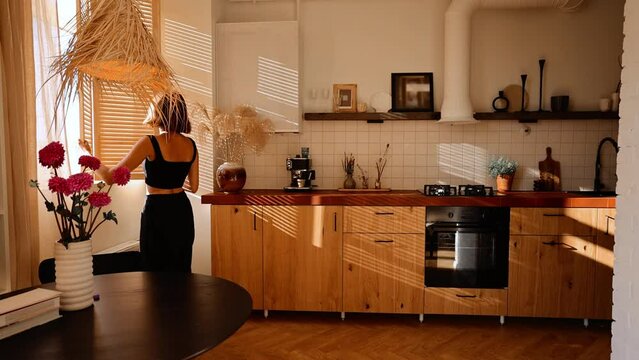 Woman opens window blinds letting the sun in and drinks a coffee, spending morning time in cozy and stylish kitchen at home