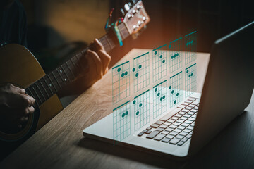 A man playing guitar in living room  while using laptop open chord on website. young guys learning music with a guitar chord table.