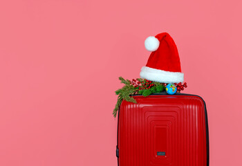 Close-up on a red suitcase is a small decorative Christmas branch, a red Santa Claus hat and a mock-up of the globe, isolate on a pink background