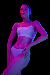 Obraz na płótnie Canvas Portrait of young beautiful woman posing in white underwear isolated over black background in neon light. Slim body