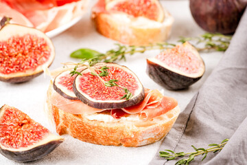 Bruschetta with raw ripe fig, cream cheese, prosciutto, walnuts and herbs on white table. Healthy quick breakfast idea. Italian cuisine. Selective focus