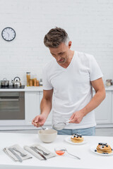 Man taking powdered sugar from bowl and holding sieve near pancakes in kitchen.