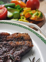 Grill Beef steak with a sprig of rosemary in a white plate. Entrecote. Rib-eye. Coarsely chopped fresh vegetables as a garnish on a wooden board - tomatoes, cucumbers, sweet peppers