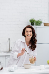 Happy brunette woman in shirt holding cereal rings near cups of coffee at home.