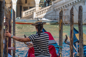 Fototapeta na wymiar The gondolier helps tourists enter the gondola near the bank of the canal of the city of Venice on a sunny morning, the Venetian gondolier extended a helping hand to the passengers of the boat