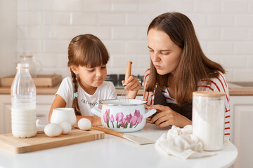 Indoor shot of woman with dark hair baking cookies or pastries with cute little preschooler girl in kitchen, sitting at table and looking in pot with dough with frowning face, disappointed with result