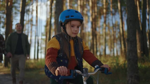 Mom and Dad teach their daughter to ride a bike on a walk in the woods. A girl rides a bicycle in a helmet