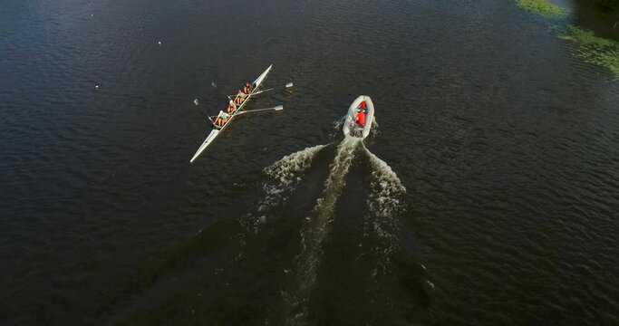 A sports kayak with a crew of four athletes sailing along the river with a coach in a motor boat, top view. Cinematic drone shot of athletes rowing in a kayak training on a calm river