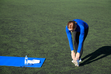 Women and sport. Girl in sportswear does exercises: bends and stretches on the grass at the stadium on a sunny day. Middle aged sportswoman dressed in sportsclothes exercising outdoors