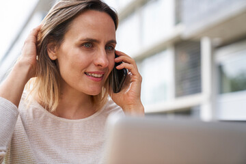 Portrait of woman sitting in front of laptop computer while speaking on cell phone