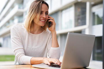 Mid-shot portrait of woman working outdoors on laptop computer while speaking on mobile phone