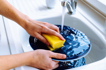 Women's hands wash dirty plate with sponge for dishes under stream of water from tap