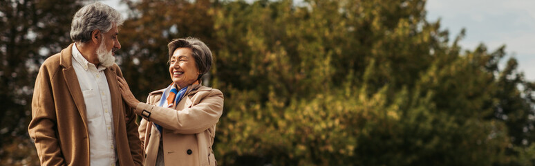 joyful senior woman hugging bearded husband in coat and standing in park, banner.