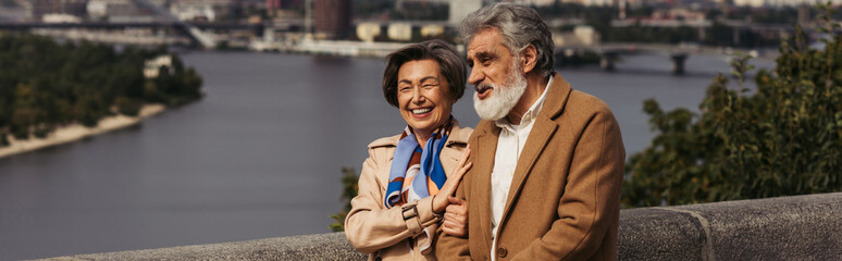 happy senior couple in beige coats smiling and walking on bridge near river, banner.
