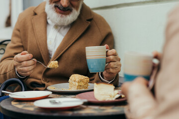 cropped view of cheerful senior man in coat holding cup of tea and eating cake near wife on terrace of cafe.