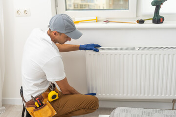 A male plumber installs a radiator in the heating system of an apartment. Guy in overalls and a gas...