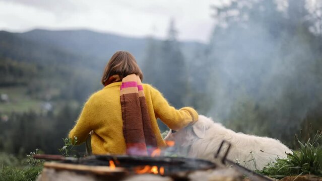 Young woman sits with her dog by the fire at campsite on nature, traveling in mountains. View from the backside. Friendship with pets and travel concept