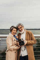 senior bearded man holding vintage camera near happy wife while standing near bridge guard rail.