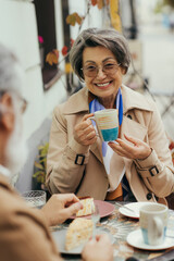 happy senior woman in eyeglasses and trench coat holding cup during brunch with husband on terrace of cafe.