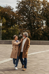 joyful senior woman hugging bearded husband in coat and standing in city park.