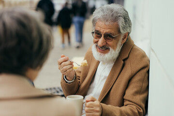 happy senior man in coat holding cup of tea and eating cake near wife on terrace of cafe.