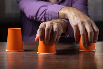 Man shows shell game of thimbles with coin, dark background. Concept deception, sleight hand.