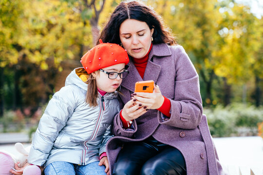Brunette caucasian woman and girl mother and daughter using smartphone sitting on the bench at city street.