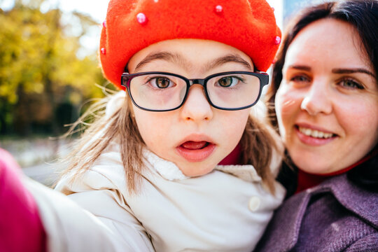 Cute curious little daughter in eyeglasses wearing red beret and mother are doing selfie outdoor in the city street. Happy family moments concept.